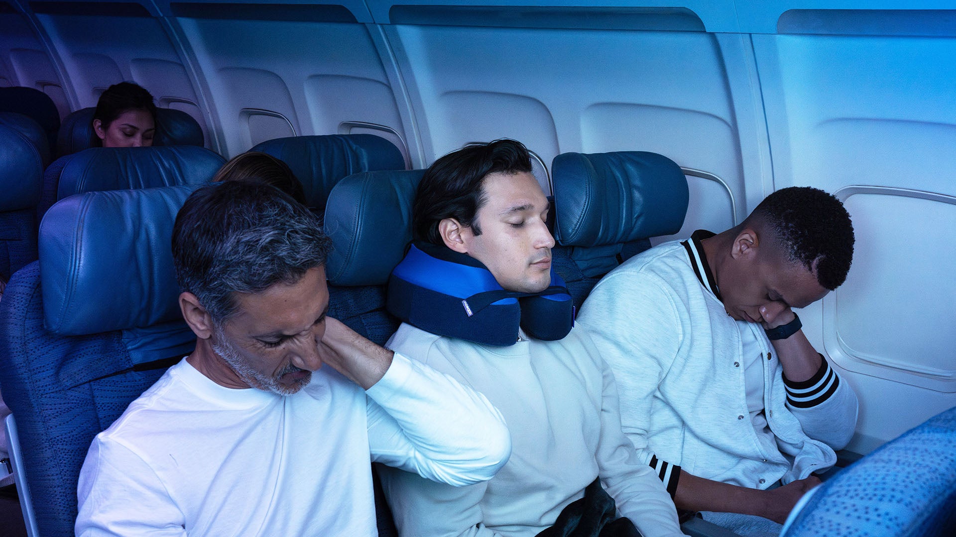 People sitting in an airplane with blue lighting, one person using a neck pillow.